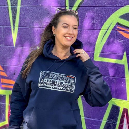 Woman in sunshine against purple graffitied wall wears Smitten silver print TB303 on navy hoodie